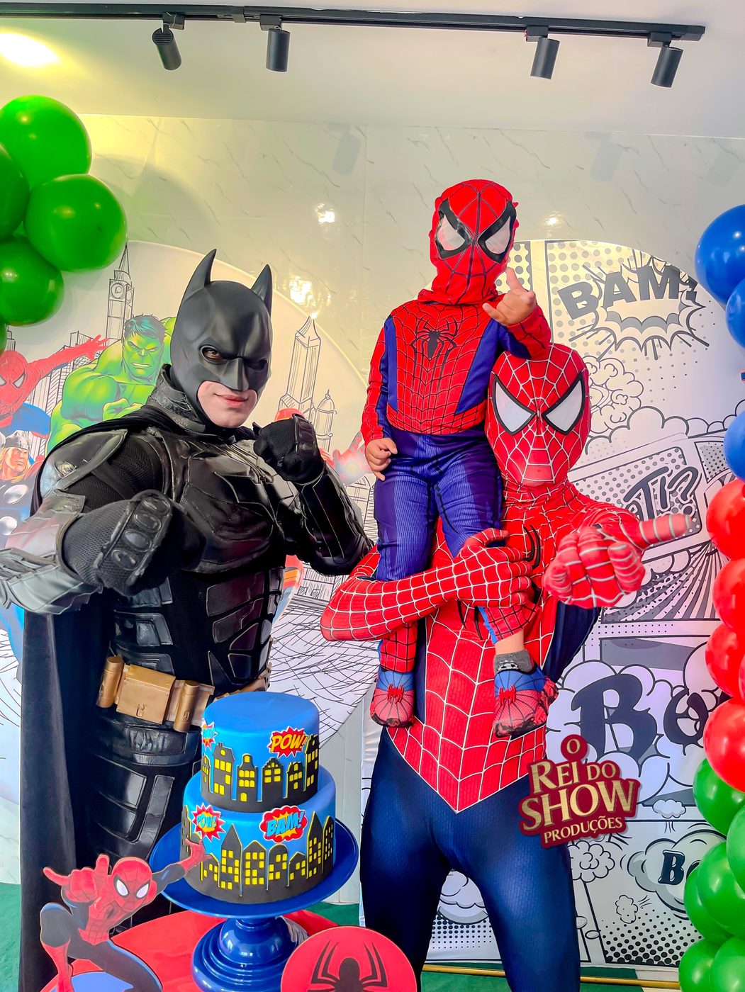 Batman e Homem-Aranha do Rei do Show com o aniversariante fantasiado, ao lado do bolo