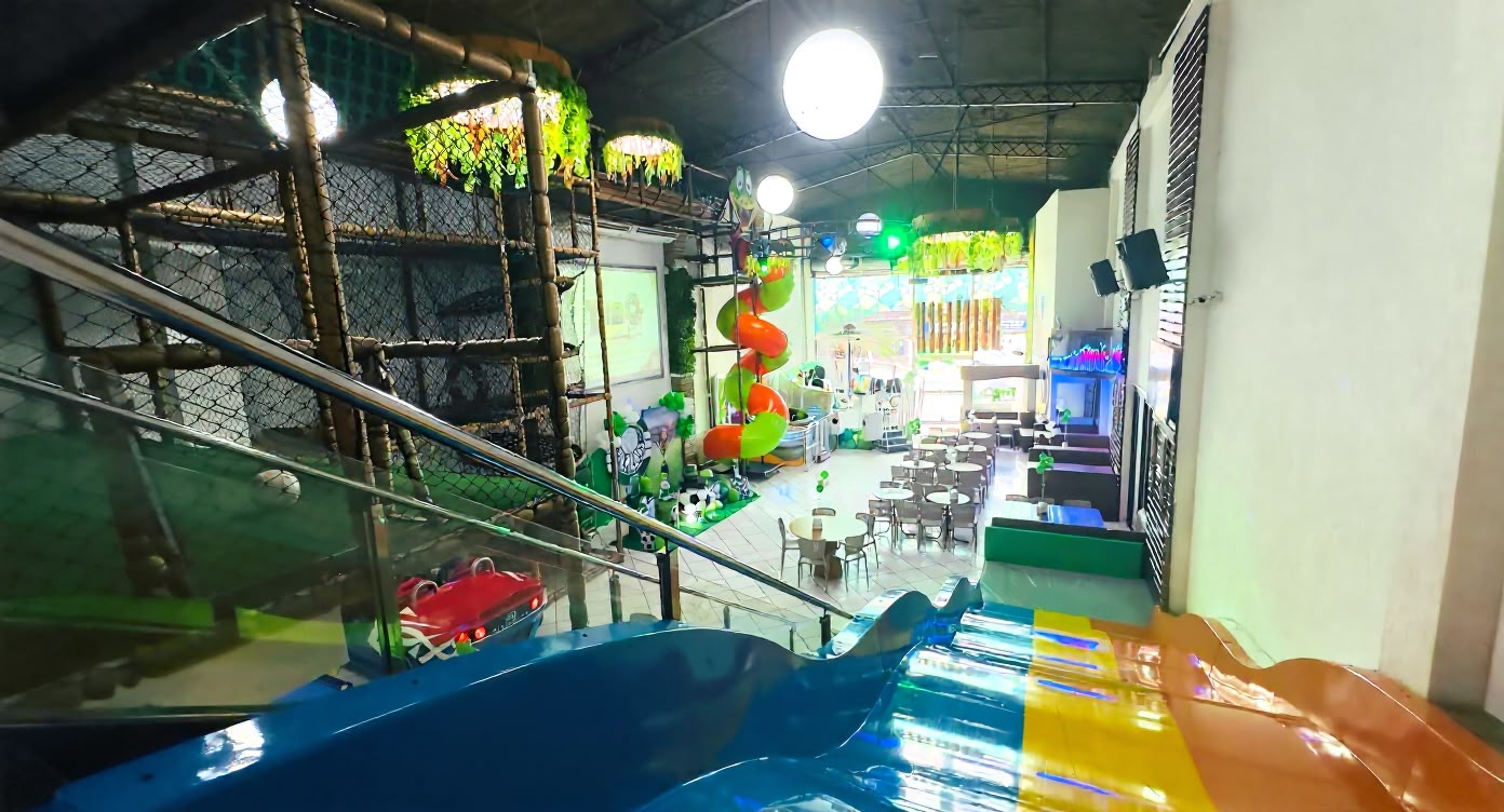 Salão da unidade Kids & Teens visto do alto do tobogã, com o brinquedão, o tobogã em espiral e as mesas